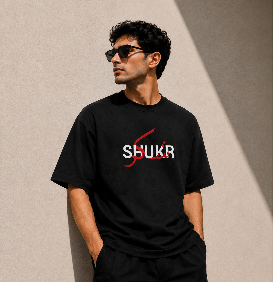 Man wearing a black t-shirt with 'SHUKR' logo against a beige wall