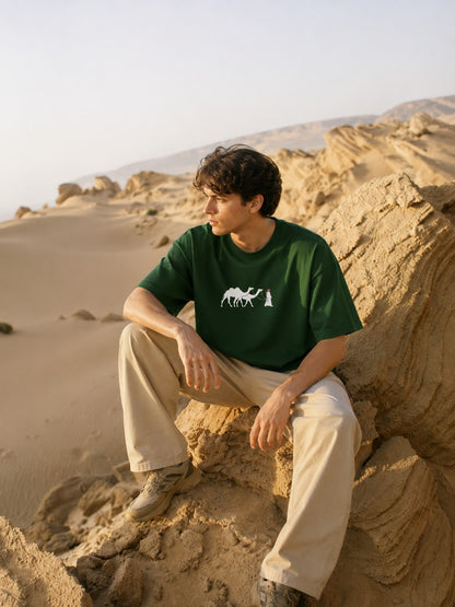 one5ive unisex oversized green cotton t-shirt with camel graphic, worn by a man sitting on desert rocks.